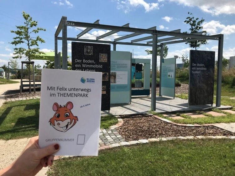 Eine Person h&auml;lt ein Natur-Rallye-Heft vor einem Outdoor-Infobereich im Themenpark hoch.