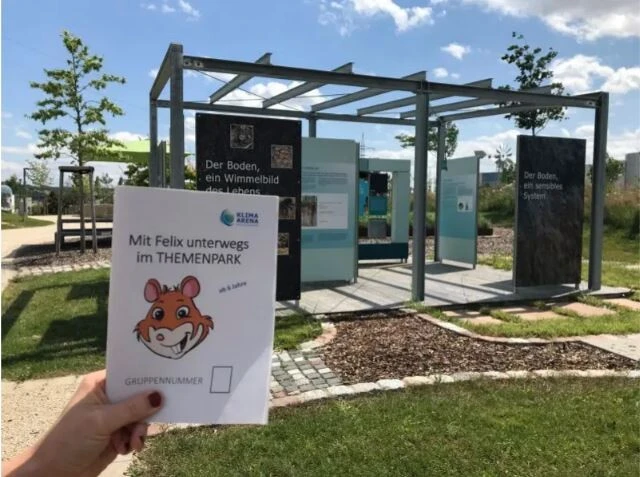 Eine Hand h&auml;lt ein Rallye-Arbeitsblatt vor einer interaktiven Outdoor-Station der KLIMA ARENA.