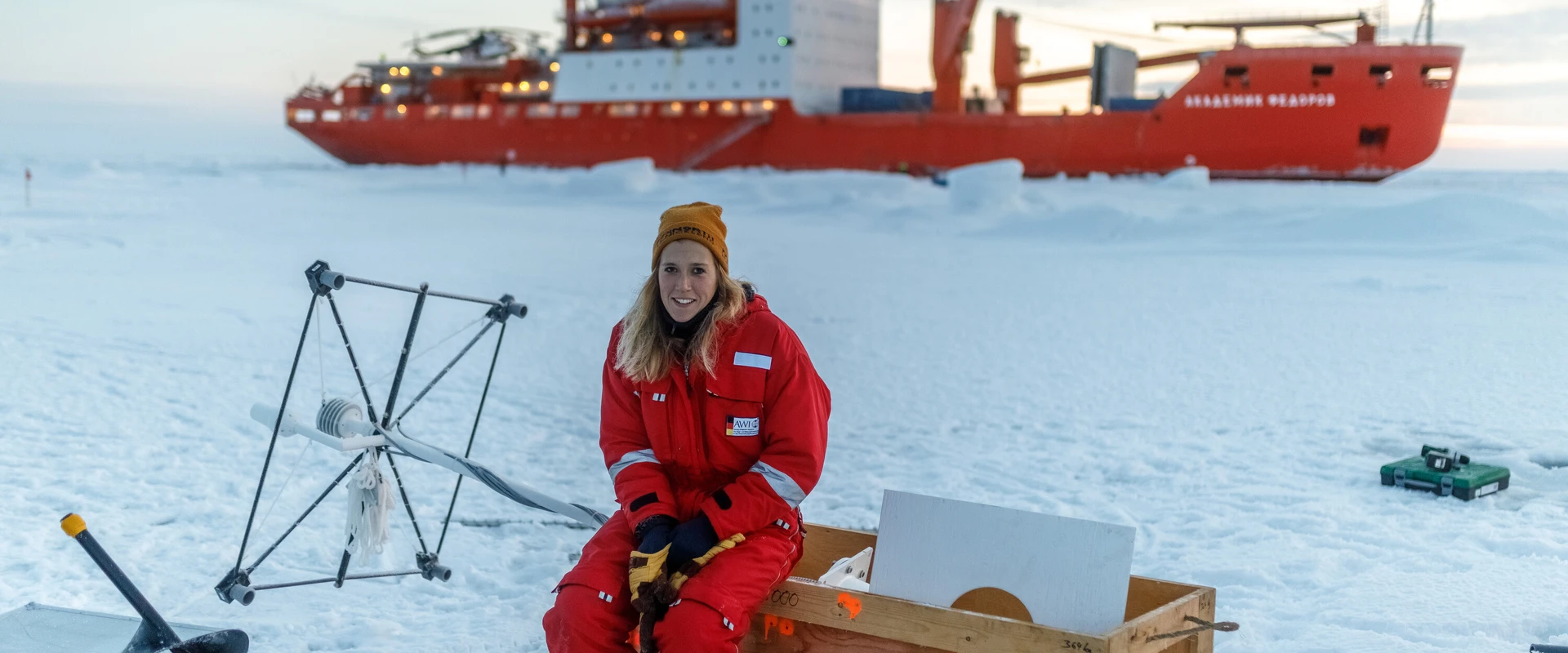 Eine Wissenschaftlerin sitzt inmitten ihrer Messger&auml;te auf einem Eisfeld. Im Hintergrund sieht man das Forschungsschiff.